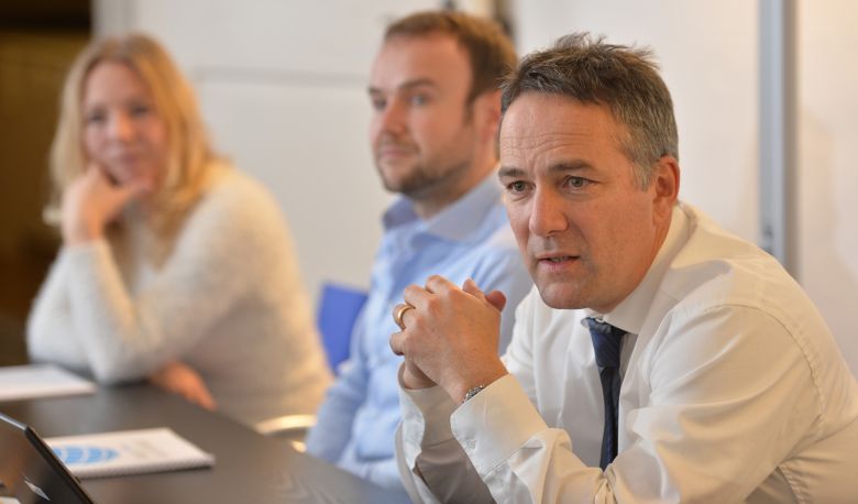Two men and a woman sit at a dark table and listen to a lecture